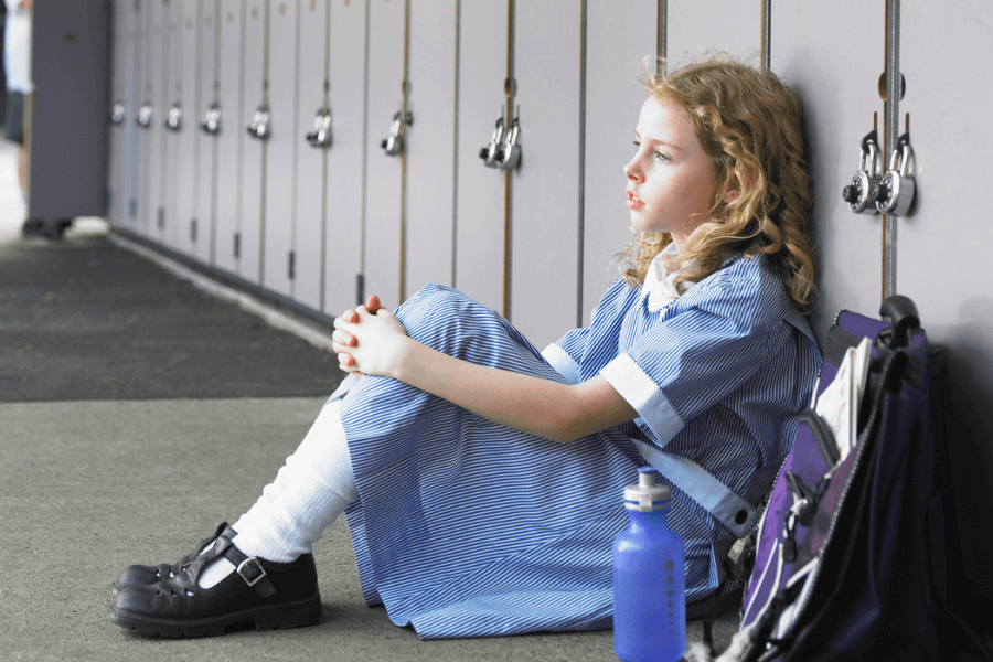 Young girl in a striped school uniform sits alone on the ground against a row of lockers, looking contemplative. Beside her are a purple backpack with books and a blue water bottle.