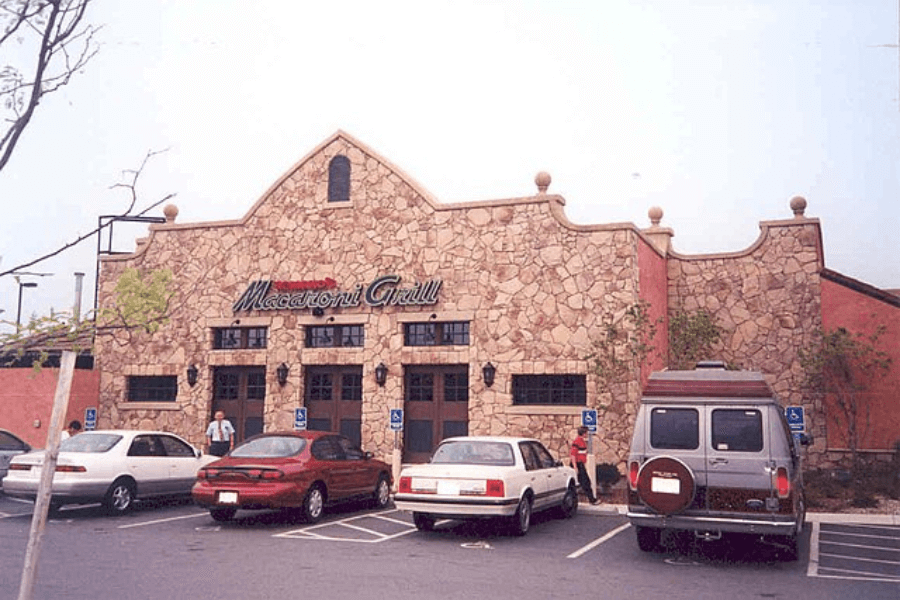 This image shows the exterior of a Macaroni Grill restaurant, featuring a rustic stone facade and arched windows, with a parking lot filled with cars, evoking an inviting, classic dining experience.