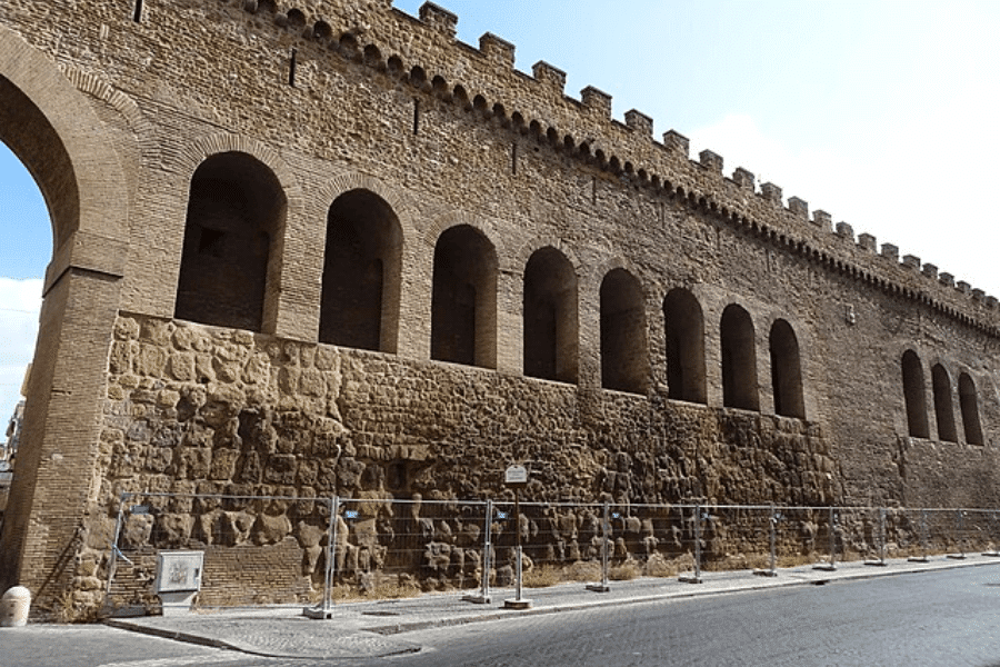 Long view of the fortified Leonine Wall in Vatican City, built with ancient stone and brickwork, featuring a series of tall arched openings and crenellated battlements, partially cordoned off with fencing along the cobbled street below.