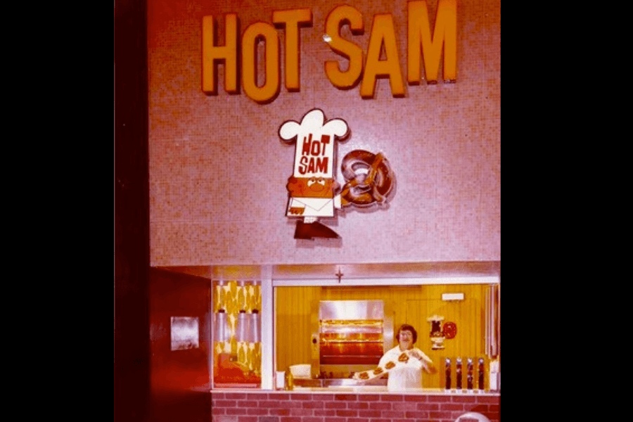 This image captures a nostalgic view of a Hot Sam pretzel stand, featuring its iconic signage with a cheerful mascot wearing a chef’s hat and a glimpse of a staff member inside the brightly lit kiosk.
