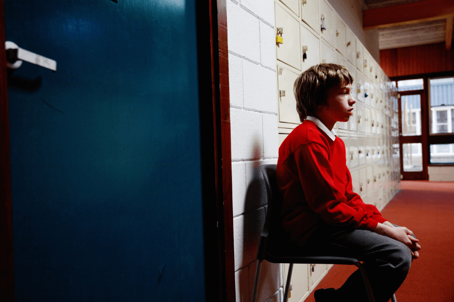 Young boy in a red sweater and gray pants sits alone on a chair in a school hallway beside a row of cream lockers and a closed blue door, appearing pensive or anxious.