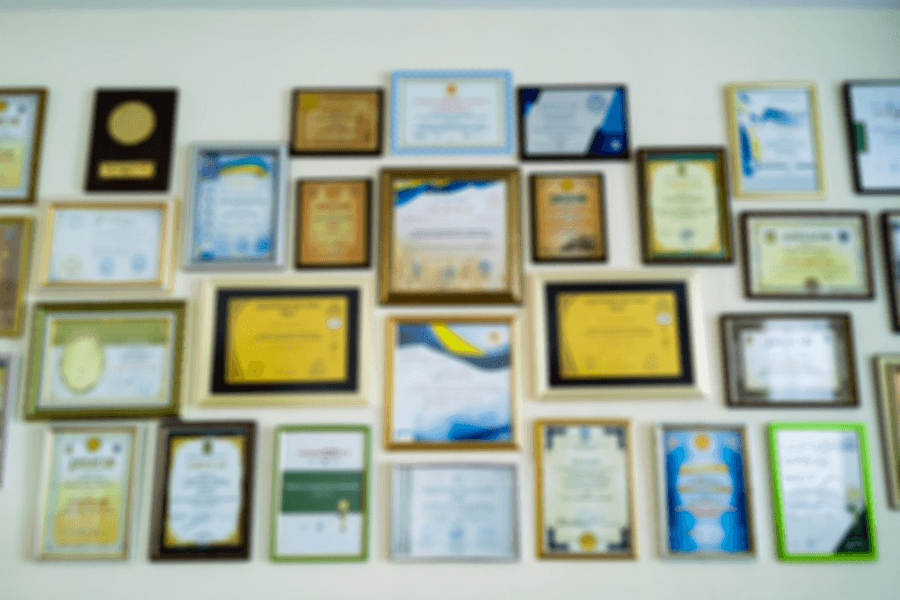 Blurred image of a wall covered in various framed certificates, awards, and diplomas, arranged in a grid pattern, suggesting recognition or achievement in an office or academic setting.