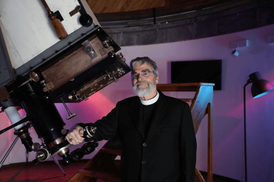 A Jesuit priest and astronomer stands beside a large telescope in an observatory room with colorful lighting, smiling as he poses for the photo; wooden stairs and a modern lamp are visible in the background.