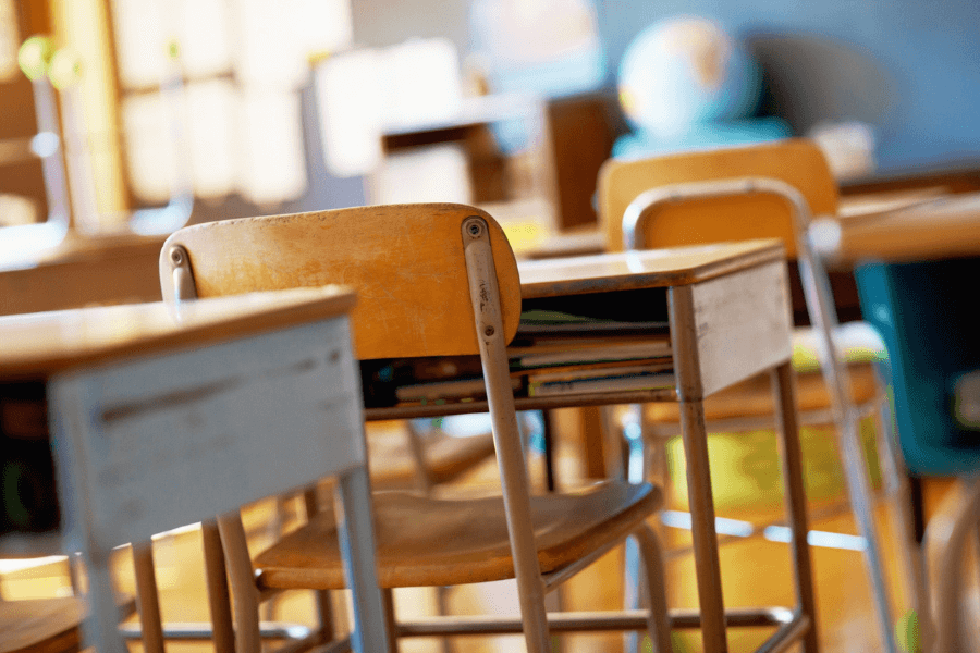 Close-up of empty wooden and metal desks and chairs in a brightly lit classroom, with a globe blurred in the background.