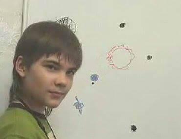 A boy with brown hair and a green shirt stands beside a whiteboard covered in hand-drawn planetary sketches and symbols, glancing toward the camera with a slight smile.