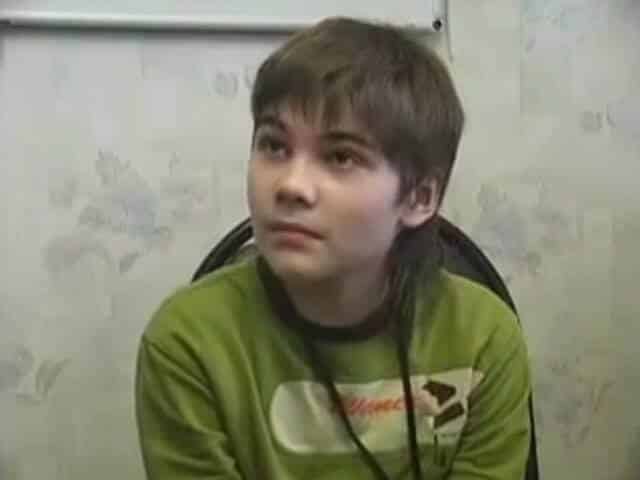 A young boy with shaggy brown hair sits on a black chair, wearing a green graphic sweatshirt and a lanyard, looking slightly upwards with a thoughtful expression.