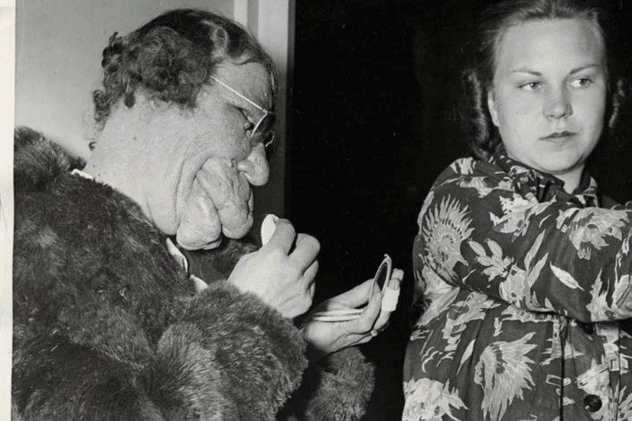 Black and white photo of two women; one on the left with a significant facial deformity is seen holding a compact mirror and applying makeup, dressed in a fur coat and glasses. The woman on the right, with a neutral expression, looks away while adjusting her sleeve. The moment captures themes of dignity, self-expression, and social contrast in the face of visible difference.