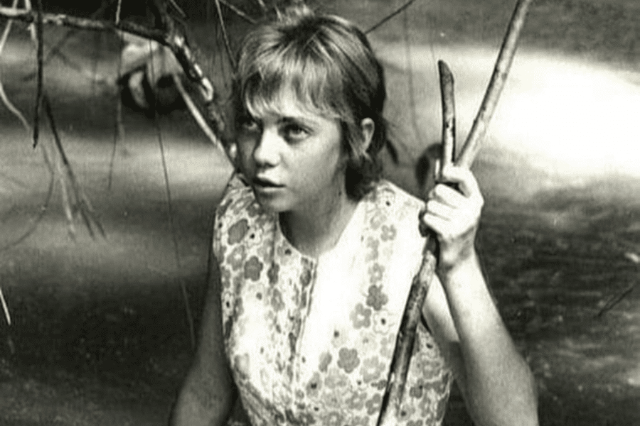 Black-and-white photo of a young girl with short hair and a floral-patterned sleeveless dress, standing in shallow water while holding onto branches. She looks alert and focused, possibly navigating through a forested or jungle area.