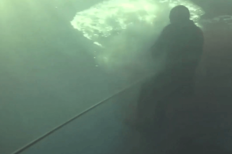 Dim underwater scene showing a diver holding onto a rope or tether near a turbulent patch of bubbles, possibly from a disturbance at the surface. The lighting is moody with limited visibility, adding an intense and suspenseful feel.