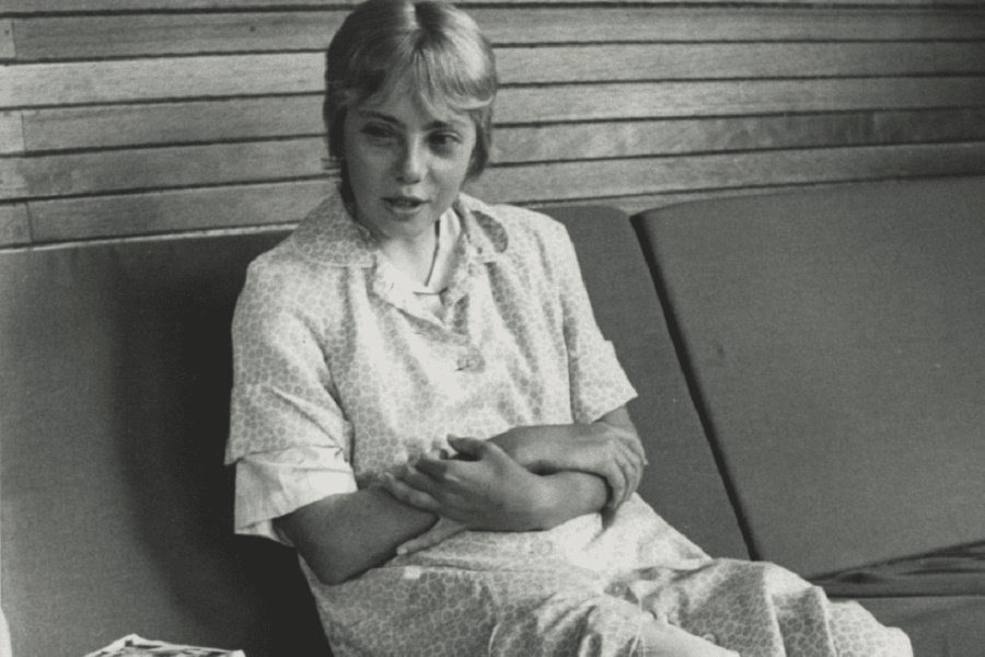Black-and-white photo of a young woman sitting on a cushioned bench with wooden paneling in the background. She wears a light-patterned dress and has her arms crossed over her midsection, appearing to be in conversation or deep thought.