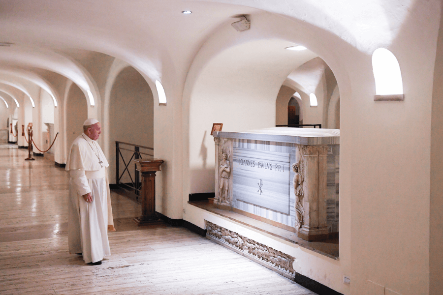 Pope Francis stands in quiet reflection before the marble tomb of Pope John Paul I in the Vatican Grottoes beneath St. Peter’s Basilica, surrounded by soft lighting and arched white corridors.