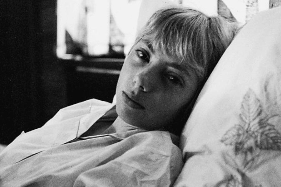 Black-and-white close-up of a young woman lying in bed, looking directly at the camera with a calm, slightly solemn expression. She is wearing a collared shirt, and a pillow with a leaf design is visible beside her.