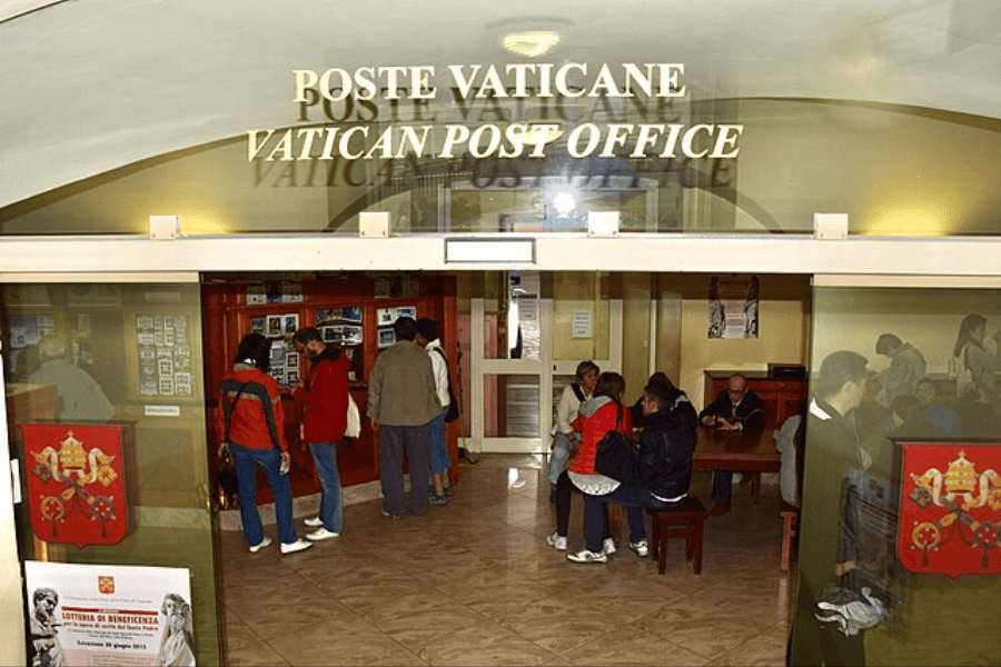 Entrance to the Vatican Post Office with a glass door displaying the sign "POSTE VATICANE – VATICAN POST OFFICE," where visitors are seen examining stamp displays and sitting at tables inside the warmly lit interior.