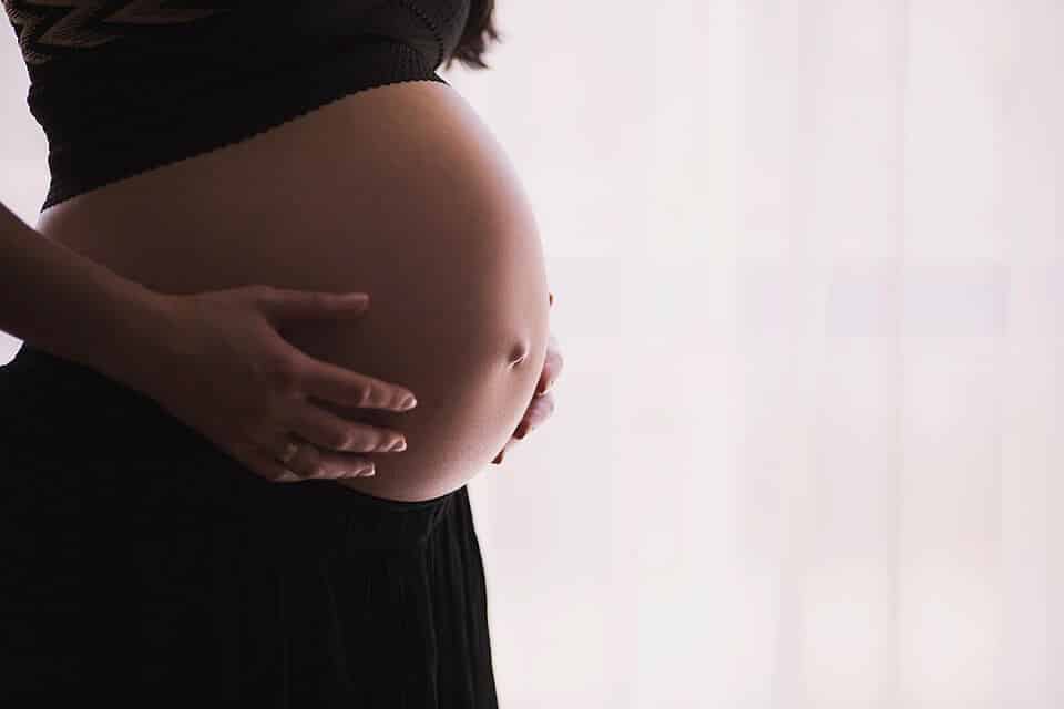 A close-up side view of a pregnant woman's bare belly, with her hands gently cradling it, standing in front of softly lit curtains.