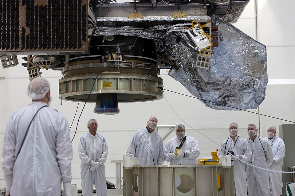 A group of engineers in white cleanroom suits and hairnets stand beneath a large, suspended spacecraft component inside a sterile laboratory, observing and monitoring the assembly process.
