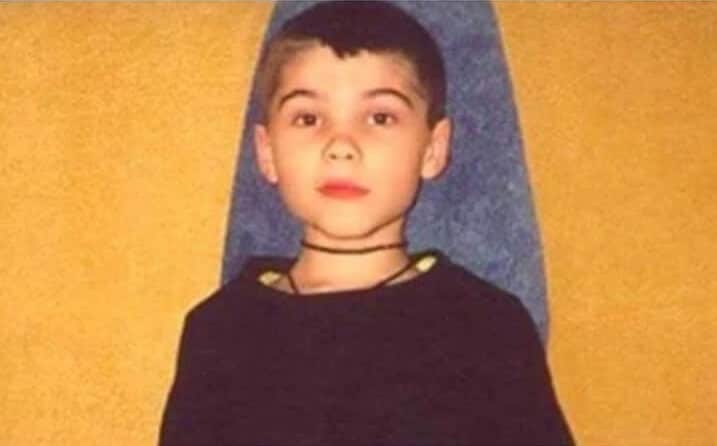 A young boy with a buzz cut and serious expression sits against a yellow and blue background, wearing a black shirt and a choker-style necklace.