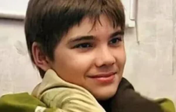 A smiling boy with straight brown hair looks to the side while someone gently places their gloved hands on his shoulders.