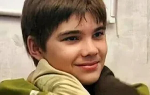 A smiling boy with straight brown hair looks to the side while someone gently places their gloved hands on his shoulders.