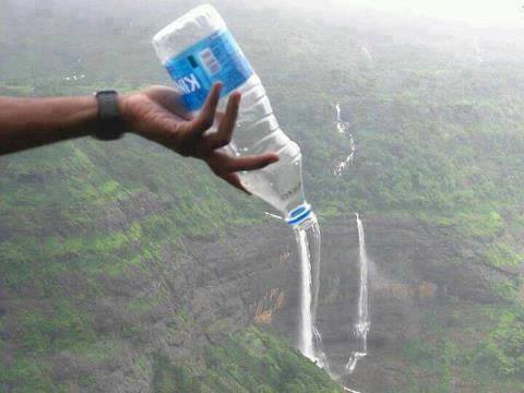 A person holds a plastic water bottle in the foreground, tilted to align perfectly with a distant waterfall in the background, creating the playful illusion that the water is pouring from the bottle. The scene is set against lush, green cliffs under misty, overcast skies.