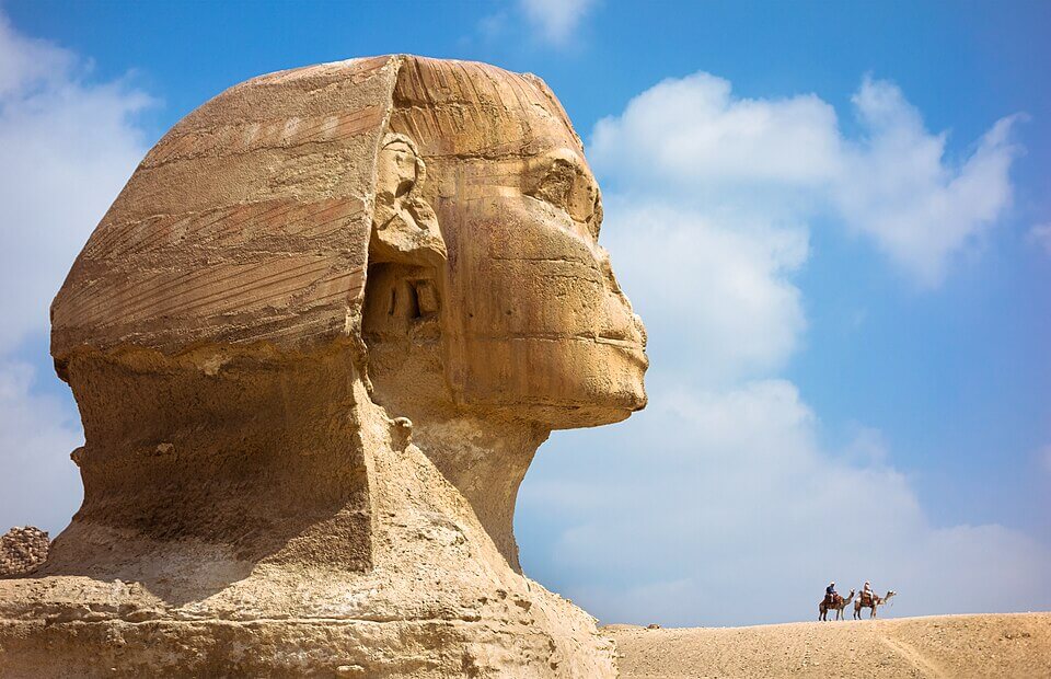 The Great Sphinx of Giza stands majestically under a bright blue sky, with two people riding camels in the distant background on the sandy plateau.