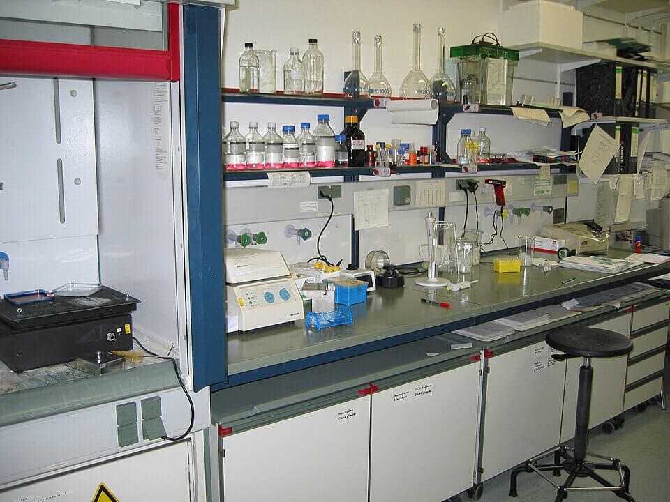 A busy science laboratory workspace with shelves full of bottles, flasks, and equipment, and a long counter holding various lab tools, notes, and a rotating stool.