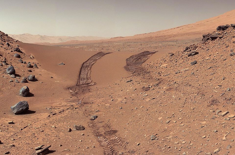 A barren, reddish-brown Martian landscape with rocky terrain and visible rover tire tracks curving through a sandy valley.