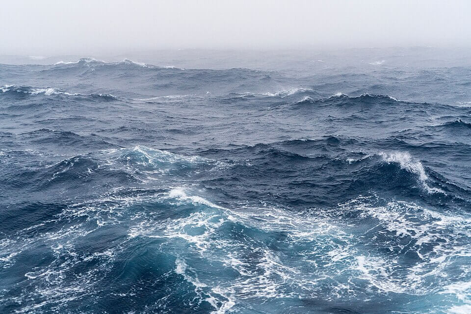 Choppy, deep blue ocean waves roll under an overcast, misty sky, with white foam forming at the crests. The horizon fades into a pale, foggy blur, evoking a cold and turbulent atmosphere.