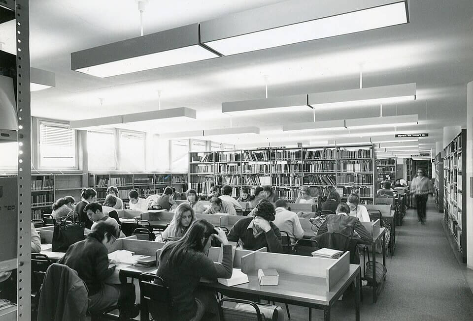 A black-and-white photo of a busy library reading room filled with students seated at study desks, surrounded by tall bookshelves and bright overhead lighting, as one person walks down the main aisle.