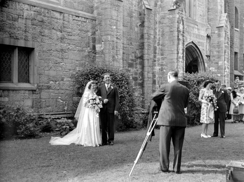 A black-and-white photo of a bride and groom posing for a wedding photo outside a stone church, with a photographer setting up a tripod and guests chatting in the background.







