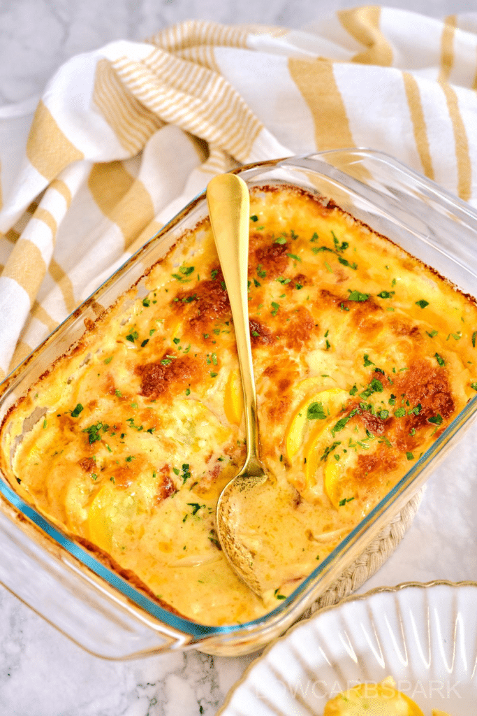 A glass baking dish filled with golden-brown, cheesy scalloped potatoes, garnished with fresh chopped parsley. A gold spoon is partially submerged, revealing the creamy, layered potato slices in a rich sauce. The dish is set on a marble countertop with a striped beige and white cloth in the background.