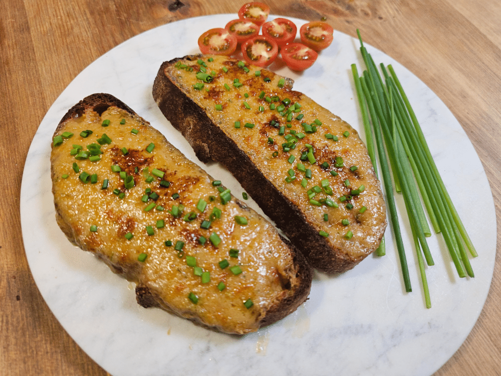 Two slices of crusty bread topped with a golden, melted cheese mixture and garnished with finely chopped chives, served on a white marble plate. The dish is accompanied by fresh cherry tomatoes sliced in half and a bundle of whole chives. The toasts have a slightly charred surface, adding a crispy texture.