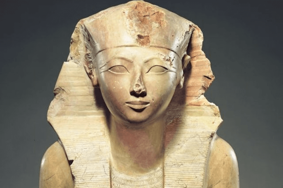 A partially damaged ancient Egyptian statue of a pharaoh, carved from stone. The figure wears a nemes headdress and has a serene expression with almond-shaped eyes and a faint smile. The sculpture is weathered, with significant damage to the nose and parts of the headdress.