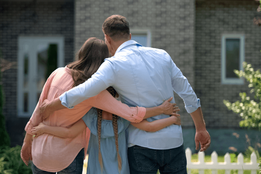 Back view of a family—two adults and a young girl with braided hair—embracing each other while standing in front of a brick house with a white picket fence, symbolizing homeownership or moving into a new home.