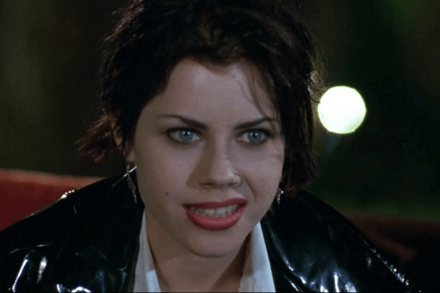This image features a woman with short, dark hair, bright eyes, and a vivid expression suggesting intensity or frustration. She is wearing a shiny black jacket over a white shirt, with an evening or dimly lit background, including blurred lights that create a dramatic atmosphere.