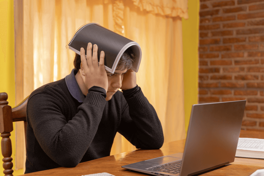 This image shows a person sitting at a desk with a laptop in front of them, appearing stressed or frustrated. They have placed an open notebook over their head as if shielding themselves or expressing exasperation. The background features warm indoor lighting with curtains and a brick wall.