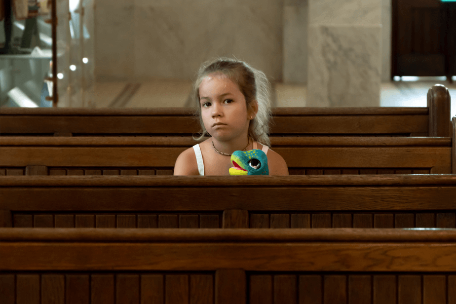 Young girl sitting alone on a wooden pew in a church, holding a colorful plush toy with a serious or contemplative expression on her face.