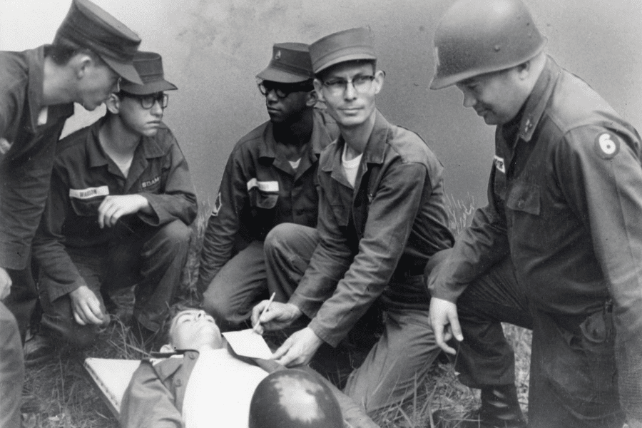 Black-and-white photo of a group of U.S. Army soldiers gathered around a medical training exercise. One soldier lies on a stretcher while others observe, as a man in glasses kneels beside the patient and writes notes on a clipboard. The team appears focused, indicating a moment of instruction or evaluation.