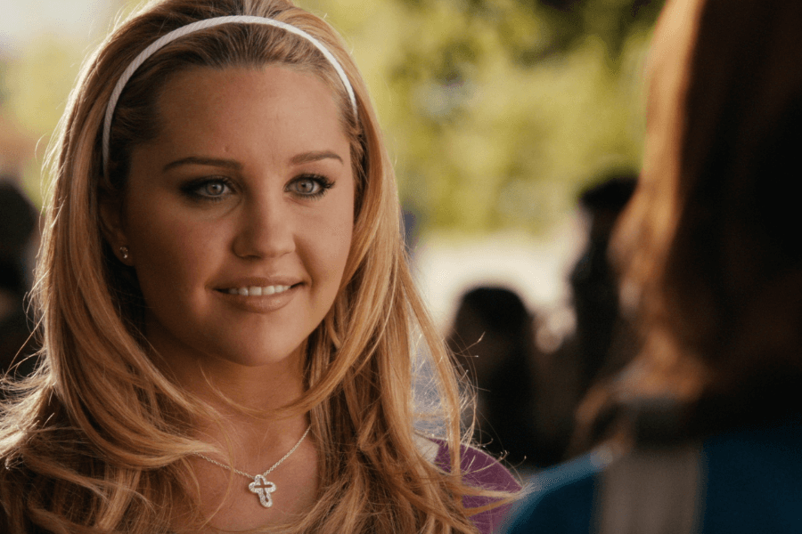 This image features a young woman with long blonde hair, a white headband, and a warm smile. She is wearing a necklace with a cross pendant, standing outdoors with a blurred background of trees and people, creating a bright and cheerful atmosphere.