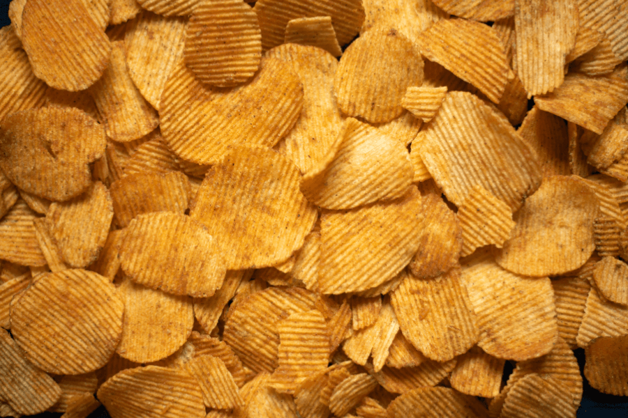 This image shows a close-up of a pile of ridged potato chips, coated with seasoning for added flavor. The chips have a golden-brown hue, highlighting their crispy texture and savory appeal.
