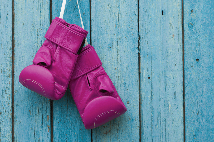 This image features a pair of bright pink boxing gloves hanging by their straps against a weathered, light blue wooden background. The contrasting colors and rustic texture create a striking and playful visual.