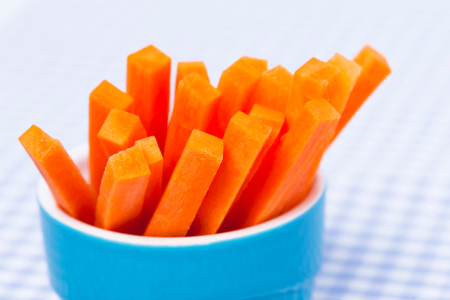 A blue cup filled with fresh, bright orange carrot sticks sits on a checkered blue and white tablecloth. The crisp texture and vibrant color of the carrots stand out against the soft background.