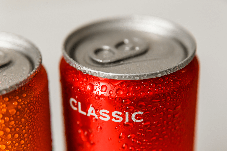 This image shows a close-up of two soda cans, with the focus on a red can labeled "Classic," covered in condensation droplets. The metallic texture and cold appearance suggest the drink is refreshing and chilled.