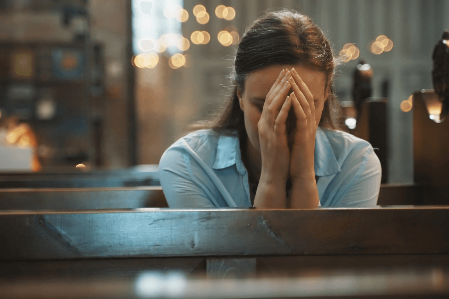 Woman sitting alone in a church pew with her head bowed and hands clasped in front of her face, appearing deep in prayer or emotional reflection, with warm, soft lighting in the background.