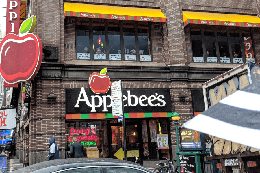 This image features the exterior of an Applebee's restaurant located in an urban setting. The building is adorned with a large red apple logo, colorful awnings, and signage advertising dining and cocktails across multiple floors. The scene also includes pedestrians, a street sign, and part of a subway entrance, giving it a bustling city atmosphere.