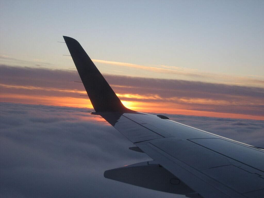 A view from an airplane window showing the wing extending into the sky, with a stunning sunset in the background. The sun casts an orange and pink glow over the clouds, creating a serene and dreamy atmosphere above the horizon.
