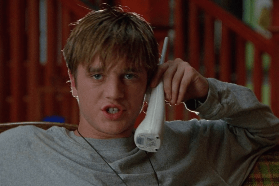 This image shows a young man with messy hair, wearing a casual gray shirt, holding a cordless phone to his ear. He appears to be mid-conversation, with a slightly expressive or animated facial expression. The background includes a warm, indoor setting with wooden elements, suggesting a relaxed, homey environment.