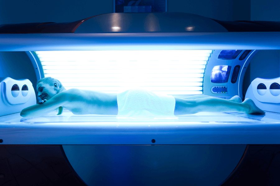 A person lies face-down in a tanning bed, illuminated by the bright blue UV lights. They are wrapped in a towel around the waist, resting comfortably with a relaxed expression.