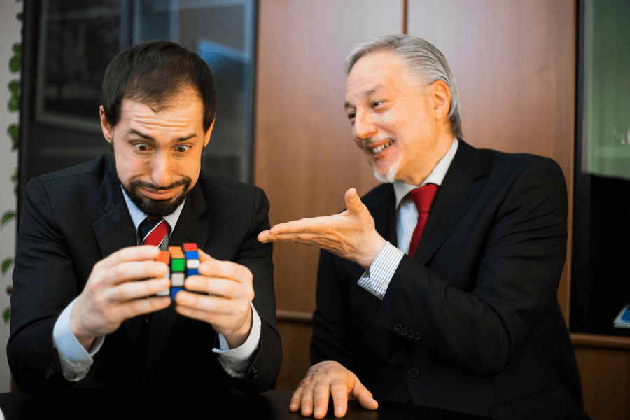 This image shows two men in business attire. One man, appearing stressed or confused, is holding and looking at a Rubik's Cube, while the other man, older and smiling, gestures toward him as if offering advice or encouragement. The setting appears to be a professional office environment, creating a humorous contrast between the puzzle and the formal atmosphere.
