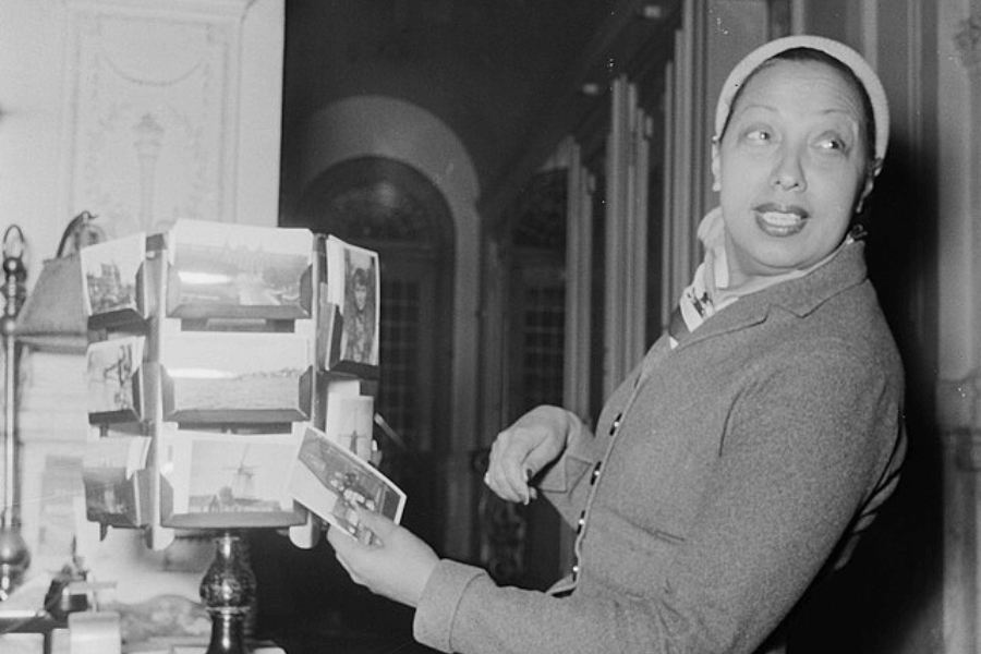 A black-and-white photograph of a woman wearing a headscarf and a buttoned-up coat, standing next to a rotating postcard rack. She holds a postcard in one hand while looking over her shoulder with an animated expression. The elegant interior setting, with arched windows and ornate decor, adds a sophisticated atmosphere.