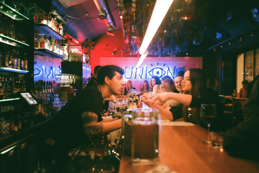 A lively bar scene with a bartender leaning over the counter to take an order from a group of customers. The background features neon lights, a graffiti-style "UNION" sign, and shelves lined with liquor bottles.